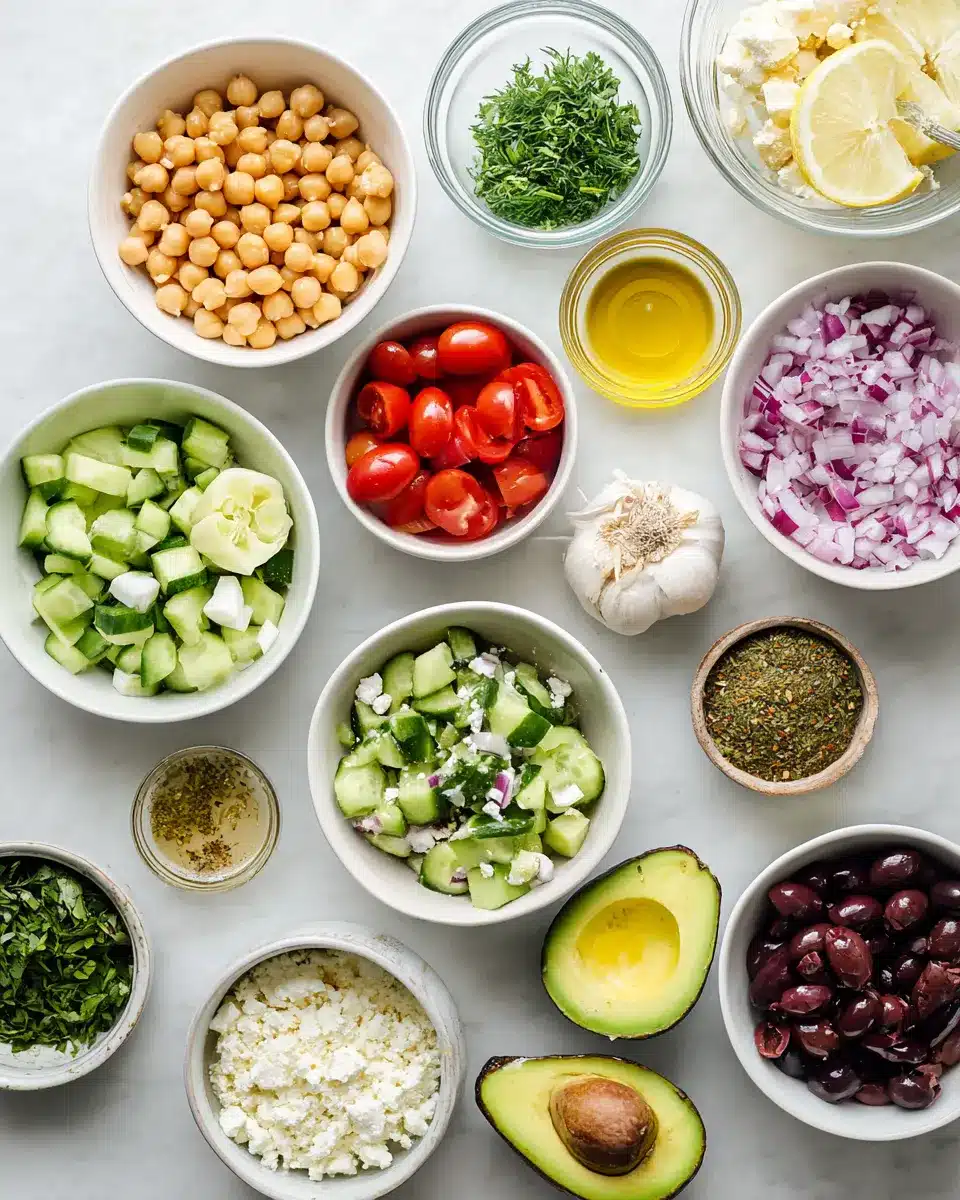 Overhead shot of chopped ingredients for chickpea and avocado salad, including avocado halves, cherry tomatoes, feta, cucumbers, and herbs.